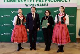 Zdjęcie przedstawia 4 osoby stojące na zielonym tle z logo UPP. Od lewej przedstawicielka zespołu Pieśni i Tańca Łany, Rektor UPP, Pani mgr Anna Binczarowska, przedstawicielka zespołu Pieśni i Tańca Łany